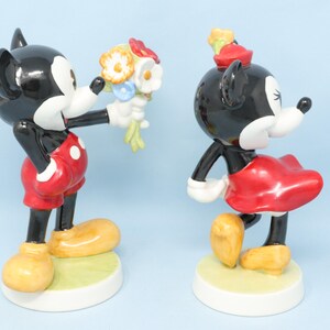 Puede incluir: Dos figuras de cer&aacute;mica de Mickey y Minnie Mouse. Mickey, con pantalones cortos rojos con lunares blancos, ofrece un ramo a Minnie, que lleva una falda y un sombrero rojos. Ambos tienen cuerpos negros, guantes blancos y zapatos amarillos.
