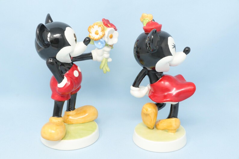 Puede incluir: Dos figuras de cer&aacute;mica de Mickey y Minnie Mouse. Mickey, con pantalones cortos rojos con lunares blancos, ofrece un ramo a Minnie, que lleva una falda y un sombrero rojos. Ambos tienen cuerpos negros, guantes blancos y zapatos amarillos.