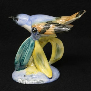 May include: A ceramic bird figurine with a blue, brown, and black body, perched on a stylized yellow and green base. The bird is in flight, with its wings spread. The base is a light blue color.