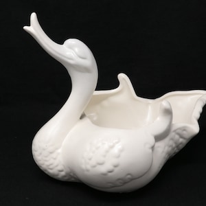 May include: White ceramic swan planter with a textured surface. The swan's neck is curved upwards and the planter has a wide, open mouth.