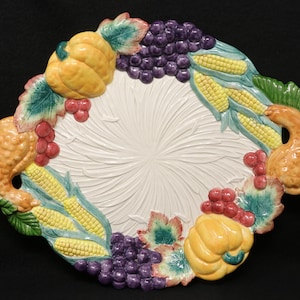 May include: A white ceramic serving platter with a raised design of fall harvest fruits and vegetables. The platter features corn, grapes, pumpkins, and leaves in shades of green, yellow, red, and purple.