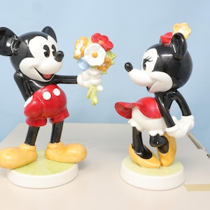 Puede incluir: Un juego de figuras de cer&aacute;mica de Mickey y Minnie Mouse. Mickey, con pantalones cortos rojos y zapatos amarillos, ofrece flores a Minnie, que lleva una falda roja y zapatos amarillos. Las figuras est&aacute;n sobre bases blancas. Colecci&oacute;n Disney.