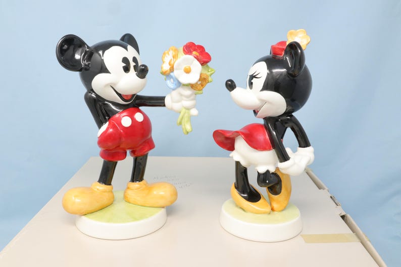 Puede incluir: Un juego de figuras de cer&aacute;mica de Mickey y Minnie Mouse. Mickey, con pantalones cortos rojos y zapatos amarillos, ofrece flores a Minnie, que lleva una falda roja y zapatos amarillos. Las figuras est&aacute;n sobre bases blancas. Colecci&oacute;n Disney.