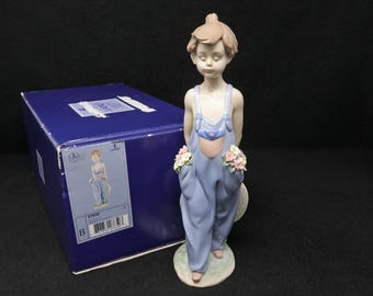May include: A porcelain figurine of a child with light brown hair wearing light blue overalls with pink and white flowers in the pockets, standing on a circular base next to a dark blue rectangular box with a product label and barcode.