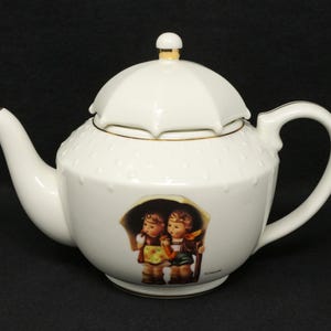 May include: White porcelain teapot with a lid and handle. The lid is shaped like an umbrella and has a gold accent. The teapot features a painted image of two children under an umbrella. The teapot has a spout and a handle.