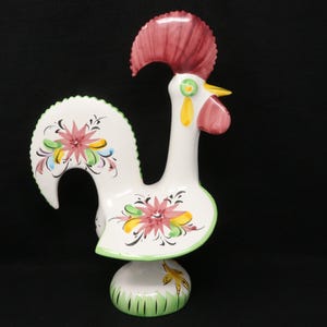 May include: A ceramic rooster figurine with a white body, a red comb, and a yellow beak. The rooster's tail and body are decorated with floral designs in pink, yellow, and green. The base is green with vertical lines.