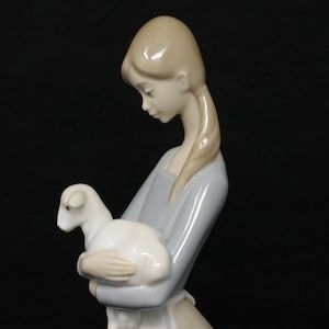 May include: A porcelain figurine of a young person holding a lamb. The figure is dressed in a light blue dress and has blonde hair. The lamb is white. The figurine is set against a black background.