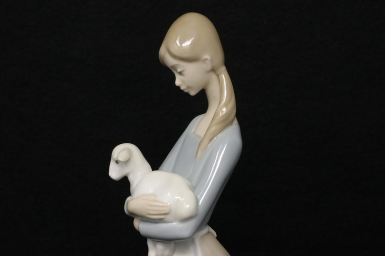 May include: A porcelain figurine of a young person holding a lamb. The figure is dressed in a light blue dress and has blonde hair. The lamb is white. The figurine is set against a black background.