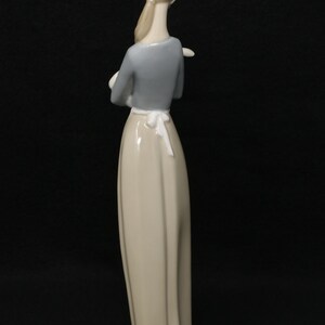 May include: A porcelain figurine of a woman, viewed from the back, with her arms crossed. She wears a light gray long-sleeved top, a long beige skirt, and a white apron tied at the waist. The figurine is on a round, light-colored base.