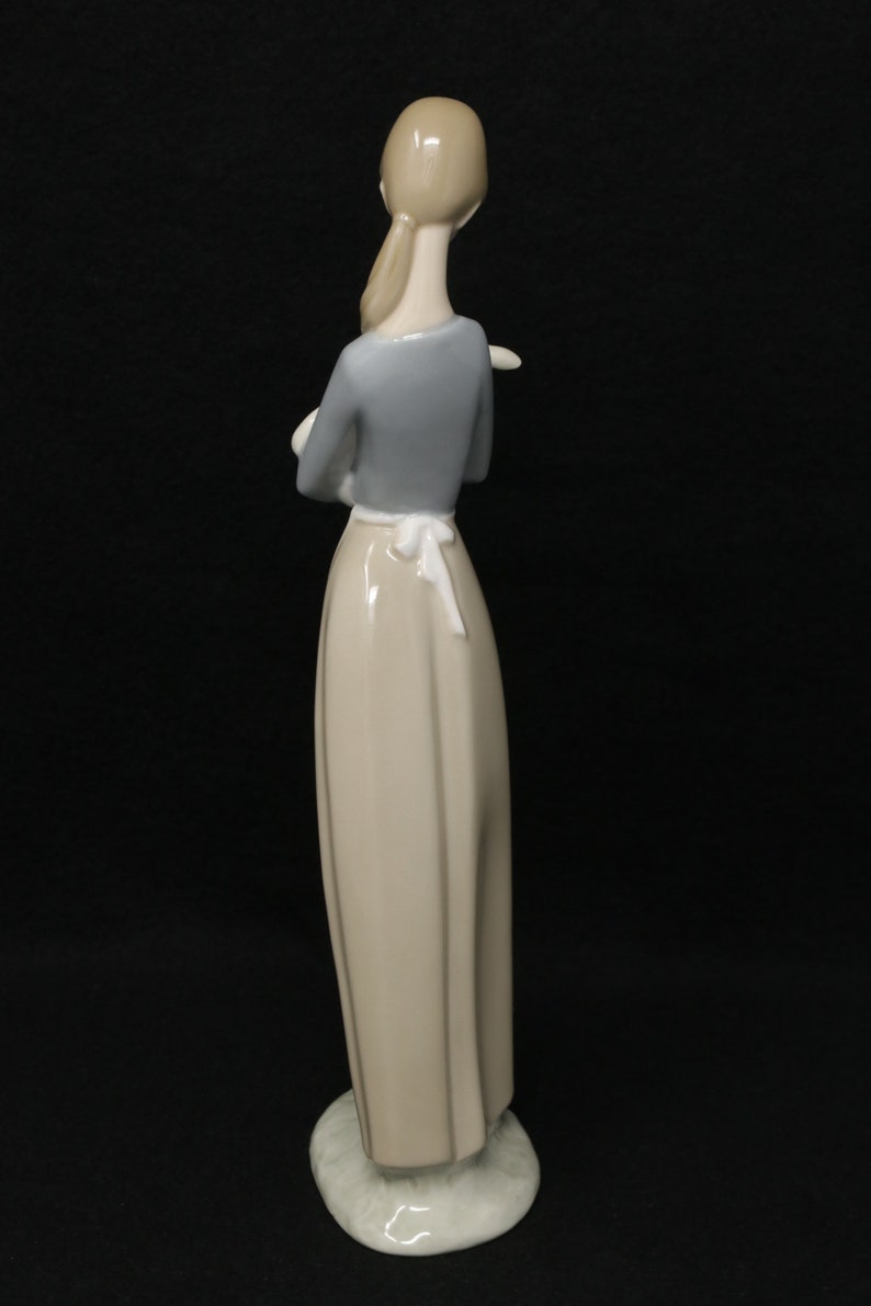 May include: A porcelain figurine of a woman, viewed from the back, with her arms crossed. She wears a light gray long-sleeved top, a long beige skirt, and a white apron tied at the waist. The figurine is on a round, light-colored base.