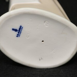 May include: Close-up of the base of a white porcelain figurine. The base is oval-shaped with a blue stamp that reads "LLADRO MADE IN SPAIN". There are also several etched markings.