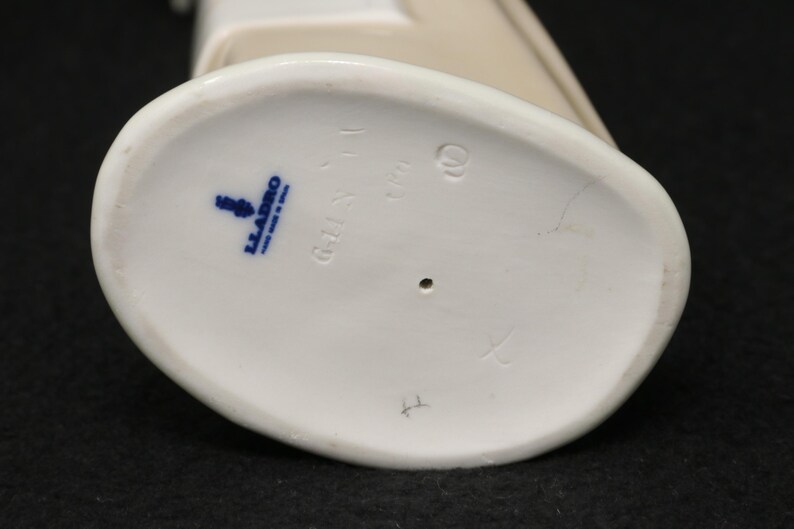 May include: Close-up of the base of a white porcelain figurine. The base is oval-shaped with a blue stamp that reads "LLADRO MADE IN SPAIN". There are also several etched markings.