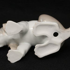 May include: A white porcelain figurine of a brown and white puppy lying on its belly with its paws outstretched. The figurine is detailed with realistic features and has a hole in the bottom for display.