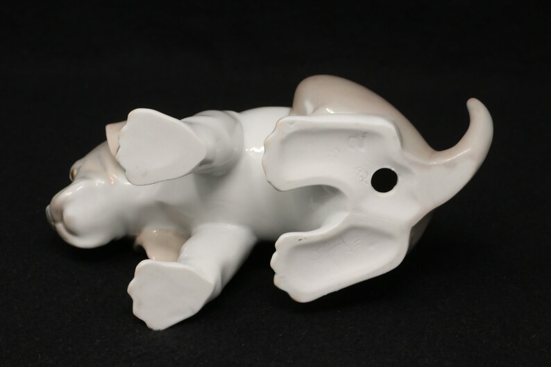 May include: A white porcelain figurine of a brown and white puppy lying on its belly with its paws outstretched. The figurine is detailed with realistic features and has a hole in the bottom for display.