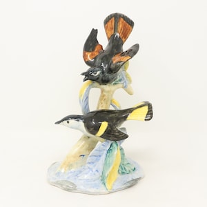 May include: A ceramic bird figurine featuring two birds perched on a branch. The birds have black and white bodies with yellow and orange accents. The base is painted with blue, yellow, and green hues.