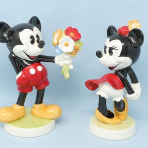 Puede incluir: Figuras de Mickey Mouse y Minnie Mouse. Mickey, con pantalones cortos rojos y zapatos amarillos, ofrece un ramo. Minnie, con falda roja y zapatos amarillos, est&aacute; cerca. Ambas figuras tienen orejas negras y guantes blancos.