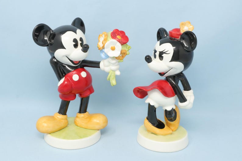 Puede incluir: Figuras de Mickey Mouse y Minnie Mouse. Mickey, con pantalones cortos rojos y zapatos amarillos, ofrece un ramo. Minnie, con falda roja y zapatos amarillos, est&aacute; cerca. Ambas figuras tienen orejas negras y guantes blancos.