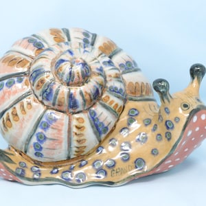 May include: Ceramic snail figurine with a detailed shell in shades of white, blue, brown, and pink. The snail's body is tan with red and white spots. The figurine has a glossy finish and is set against a light blue background.