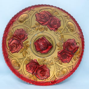 May include: A round, decorative tray with a red and gold floral design. The tray features embossed red roses and gold accents, with a scalloped red edge. The tray is approximately 12 inches in diameter.