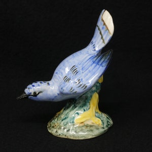 May include: A blue ceramic bird figurine perched on a green and yellow base. The bird has a long, thin beak and its wings are spread out.