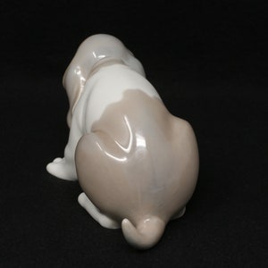 May include: A porcelain figurine of a brown and white basset hound puppy sitting with its tail curled. The puppy is looking to the left.