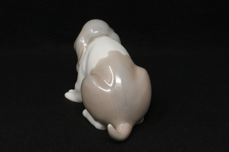 May include: A porcelain figurine of a brown and white basset hound puppy sitting with its tail curled. The puppy is looking to the left.