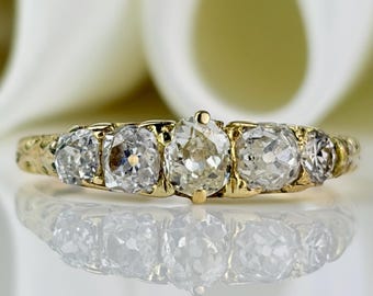 Può includere: Un anello d'oro vintage con cinque diamanti chiari e scintillanti. I diamanti sono incastonati in una fascia d'oro dettagliata, con il diamante centrale leggermente più grande. L'anello è esposto su una superficie riflettente.
