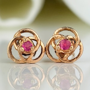 May include: A pair of gold-toned stud earrings. Each earring features a central, round, bright pink gemstone surrounded by a swirling, openwork design. The earrings are displayed on a reflective surface, with a soft, blurred background.