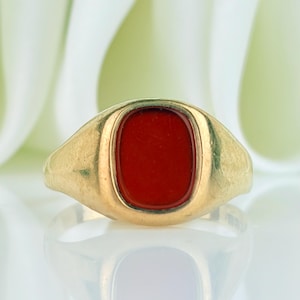 Puede incluir: Un anillo de sello de oro con una incrustación de piedra roja rectangular y lisa en el centro de la banda. El metal tiene un acabado pulido y la piedra está al ras con el engaste de oro circundante, creando un perfil de superficie uniforme.
