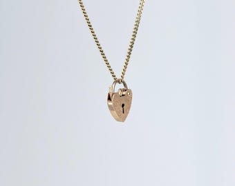 May include: A gold-colored necklace featuring a heart-shaped pendant with a keyhole design. The pendant hangs from a delicate chain. The necklace is displayed against a plain white background, highlighting the jewelry's details.