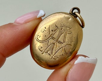Könnte beinhalten: Ein ovales Goldmedaillon mit einem gravierten Monogramm. Das Medaillon hat oben eine Schlaufe zum Anbringen einer Kette.