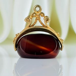 Peut inclure: Un pendentif doré avec une grosse pierre ovale brun-rouge. Le pendentif a un dessus orné et décoratif avec une petite boucle pour une chaîne. La pierre a une finition polie et brillante. Le pendentif est sur un fond flou.