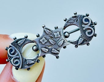May include: An antique silver brooch with a star and two crescent moon shapes. The brooch has detailed floral and scrollwork, with a pin closure. The brooch is held by a person's fingers.