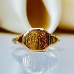 Könnte beinhalten: Ein goldener Siegelring mit einem eingravierten Monogramm. Der Ring hat eine rechteckige Oberfläche mit abgerundeten Ecken und eine polierte Oberfläche. Der Ring ist vor einem weichen, cremefarbenen Hintergrund platziert.