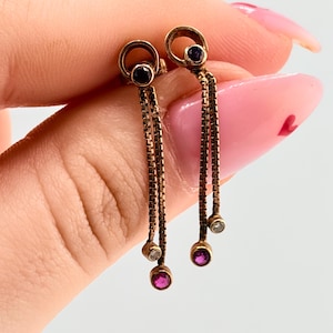 Peut inclure: Boucles d'oreilles pendantes dorées avec un dessus circulaire et des détails de chaîne. Chaque boucle d'oreille comporte une gemme violet foncé en haut et une gemme plus petite en bas, avec une gemme claire entre les deux. Sur fond clair.