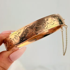 May include: A gold-coloured bangle bracelet with an engraved floral design. The hinged bracelet has a safety chain. The bracelet is held by a hand with light pink nail polish.