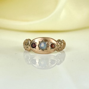 Può includere: Un anello dorato con un elemento centrale ovale. L'anello presenta una gemma centrale di colore chiaro, affiancata da due gemme rosse più piccole. Intricati riccioli adornano le spalle dell'anello. L'anello è esposto su una superficie riflettente.