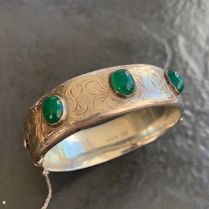 Könnte beinhalten: Ein silbernes Armreif-Armband mit einem gravierten Design und drei grünen ovalen Steinen. Das Armband hat einen Scharnierverschluss und eine Kette.