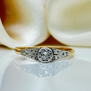 May include: A gold ring with a central diamond and smaller accent stones. The ring features a detailed design around the main stone and along the band. The ring is set against a soft, off-white background.