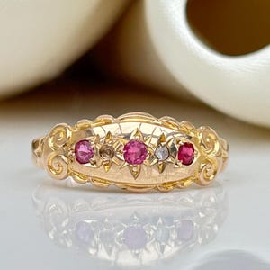 May include: A gold ring with a delicate design featuring a row of five gemstones. The ring has a central diamond and four ruby stones.