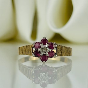May include: A gold ring with a flower design, set with six deep red gemstones and a central cluster of small diamonds. The band has a textured, ridged pattern. The ring is shown on a reflective surface.