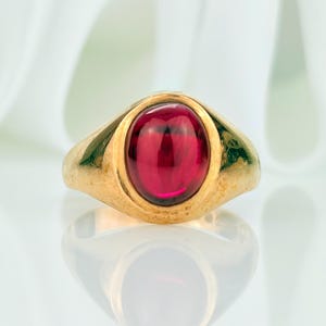 May include: A gold ring featuring a large, oval, deep red gemstone. The ring has a classic design with a smooth, polished band and a bezel setting. The ring is displayed on a reflective surface, with its reflection visible.