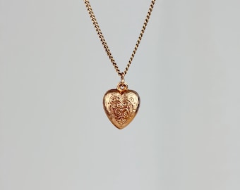 May include: A gold-toned metal necklace featuring a delicate curb chain and a heart-shaped locket pendant. The locket surface is decorated with an intricate, symmetrical embossed scrollwork pattern across the front, suspended from a small jump ring.