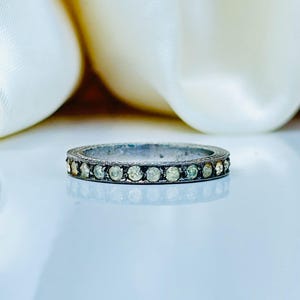 Op de afbeelding: Een zilverkleurige ring met een band van kleine, ronde, lichtgekleurde stenen. De ring is geplaatst tegen een zachte, witte achtergrond, die het delicate ontwerp benadrukt. De stenen zijn gelijkmatig verdeeld over de band.