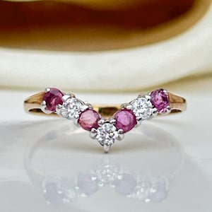 Pode incluir: Um anel de banda de ouro com uma configuração em forma de V com pedras preciosas rosa e brancas. O anel está sobre um fundo branco.