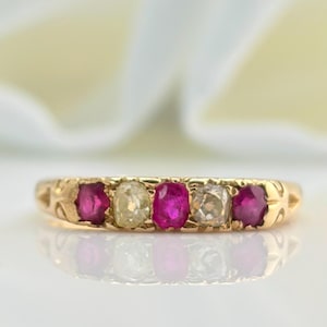 Może przedstawiać: Złoty pierścionek z naprzemiennymi czerwonymi i przezroczystymi kamieniami szlachetnymi. Pierścionek ma delikatny wzór z rzędem pięciu kamieni. Kamienie szlachetne są prostokątne i osadzone w złotej oprawie.