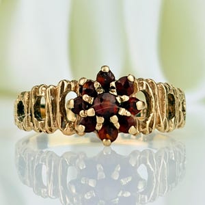 May include: A gold ring featuring a central cluster of seven round, deep red garnet gemstones arranged in a flower-like pattern. The band has a textured, vertical ribbed design that extends around the finger, providing a distinct metallic finish.