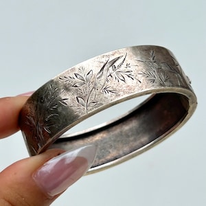 May include: Silver hinged bangle bracelet with an etched floral and bird design. The bracelet has a rectangular shape and a clasp closure. The etched details include leaves and a bird. The bracelet is held by a hand.