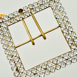 May include: A gold-toned metal buckle with a square frame, adorned with a double row of clear rhinestones.