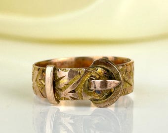 May include: A vintage gold-tone ring with a buckle design. The ring features intricate floral and leaf patterns, with a buckle clasp detail. The ring's surface shows signs of wear, adding to its antique charm. The ring is a unique piece of jewelry.
